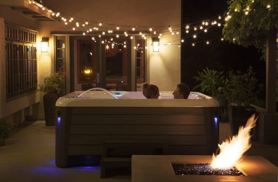 a couple enjoying a lovely evening in a highlife aria NXT spa with a lit firepit in the front and twinkling tiny patio lights overhead