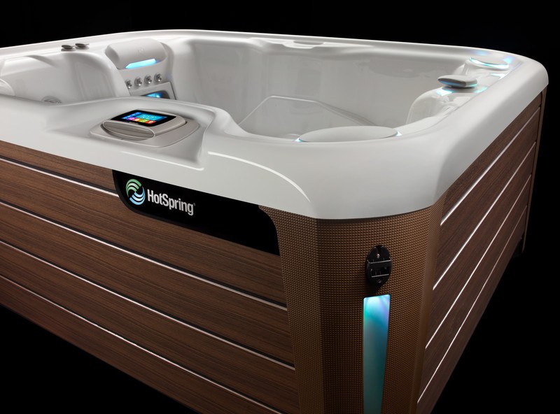 A sleek and modern Hot Spring spa with a stylish wood-paneled exterior and a smooth, contoured white shell. The spa features ambient LED lighting, an advanced touchscreen control panel, and ergonomic seating.