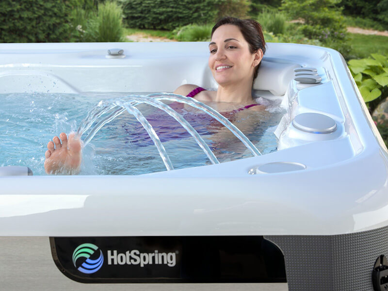 Smiling woman relaxing in a HotSpring spa, enjoying the warm water and arcing jet streams, surrounded by lush greenery.