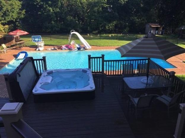 backyard view of a swimming pool with a slide, surrounded by a deck featuring a hot tub, patio furniture, and a large umbrella, with lush greenery in the background.